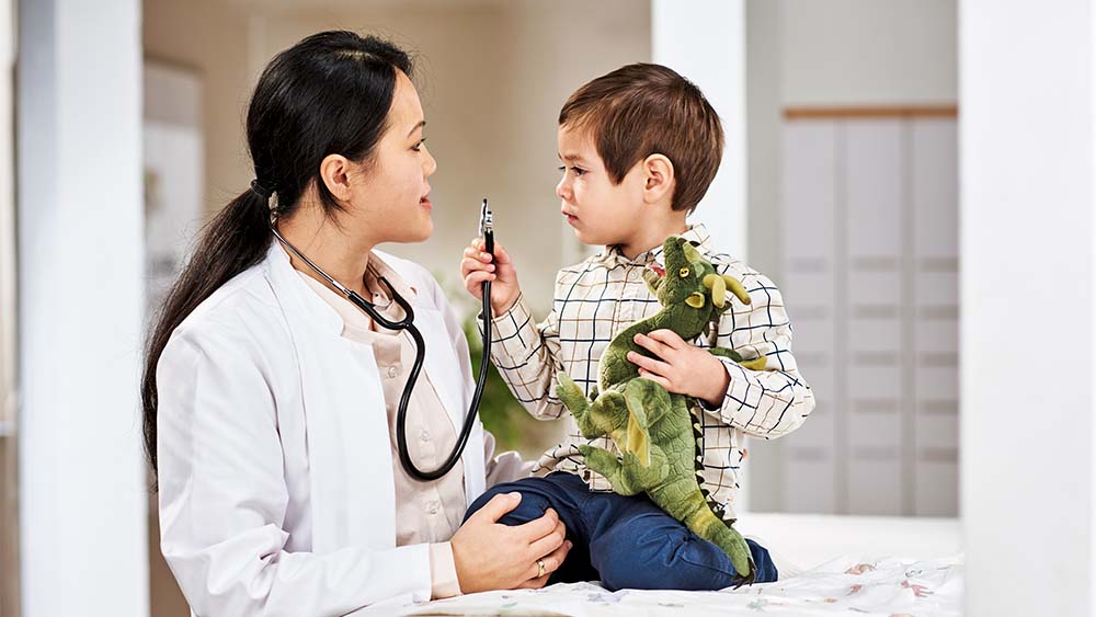 Một chuyên gia y tế nữ đang trò chuyện với một bé trai đang cầm con khủng long đồ chơi. Cậu bé trông gắn bó và tò mò, còn chuyên gia y tế thể hiện sự ấm áp và thân thiện