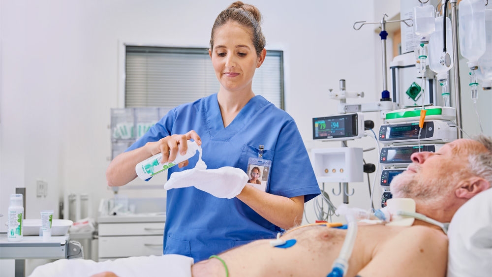 nữ y tá cầm chai Prontoderm trong bệnh viện trước một bệnh nhân cao tuổi nằm trên giường và dùng găng thoa sản phẩm
