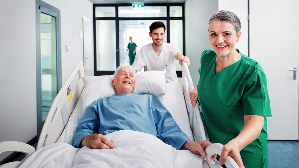 Bệnh nhân nằm trên giường bệnh được đội ngũ nhân viên y tế di chuyển qua hành lang bệnh viện, với một nhân viên khác đi cùng hỗ trợ