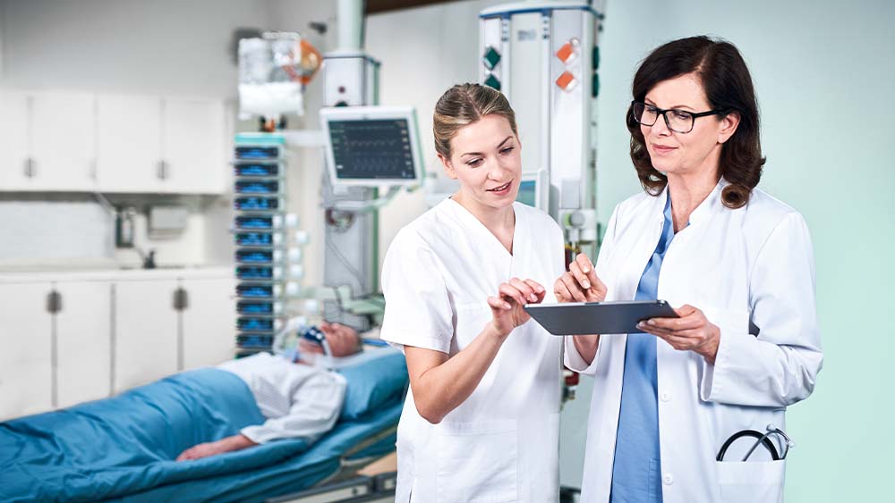 Một bác sĩ và một y tá đang thảo luận về liều lượng truyền dịch thông minh B. Braun đang được nhập trực tiếp từ máy tính bảng. Bệnh nhân trong phòng ICU có thể được nhìn thấy trong nền, được kết nối với một tháp chứa đầy các thiết bị truyền dịch thông minh.