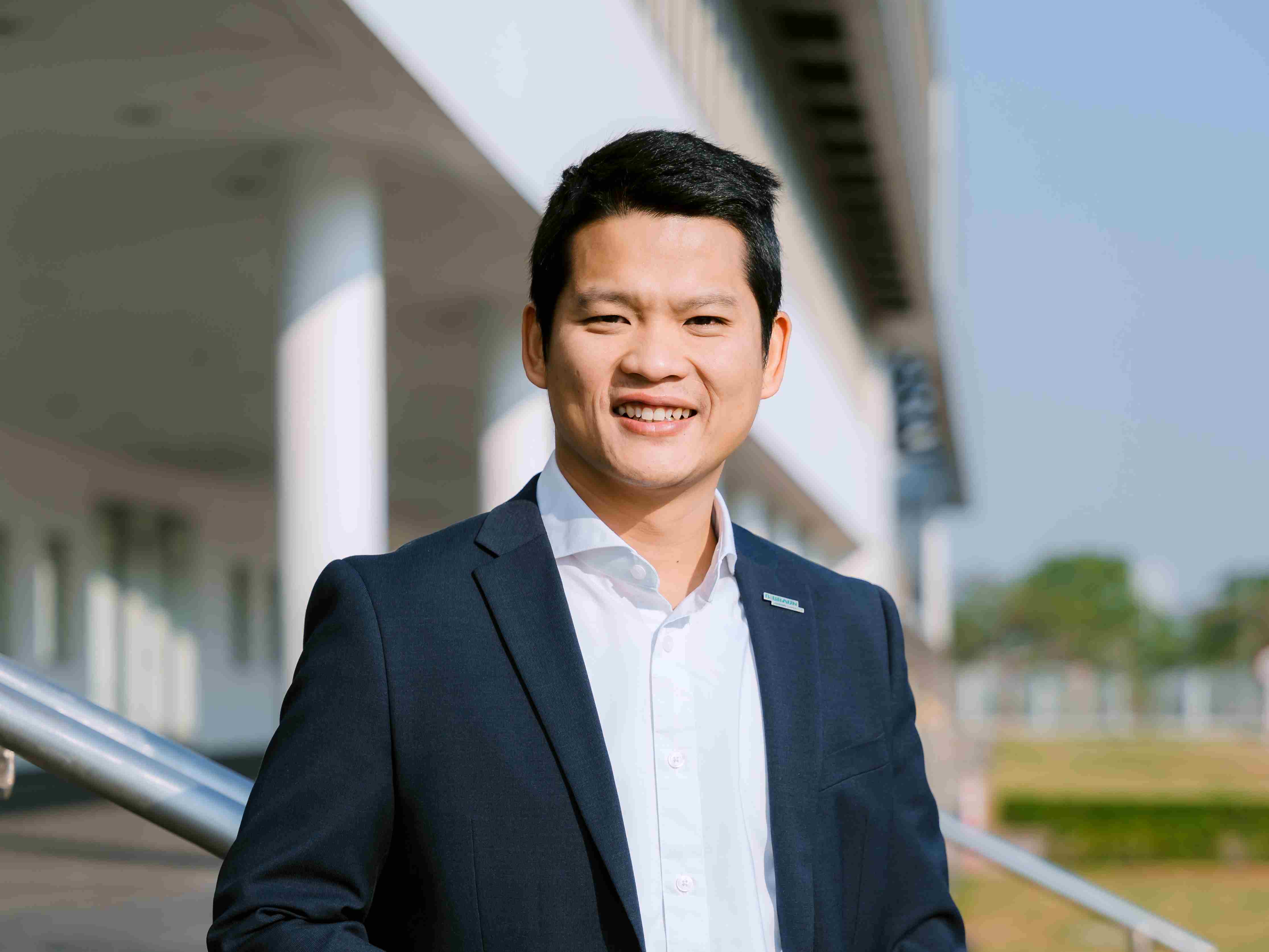 Cận cảnh một người đang cười tươi, mặc áo màu vàng, đeo kính và đứng trong khung cảnh tươi sáng. 