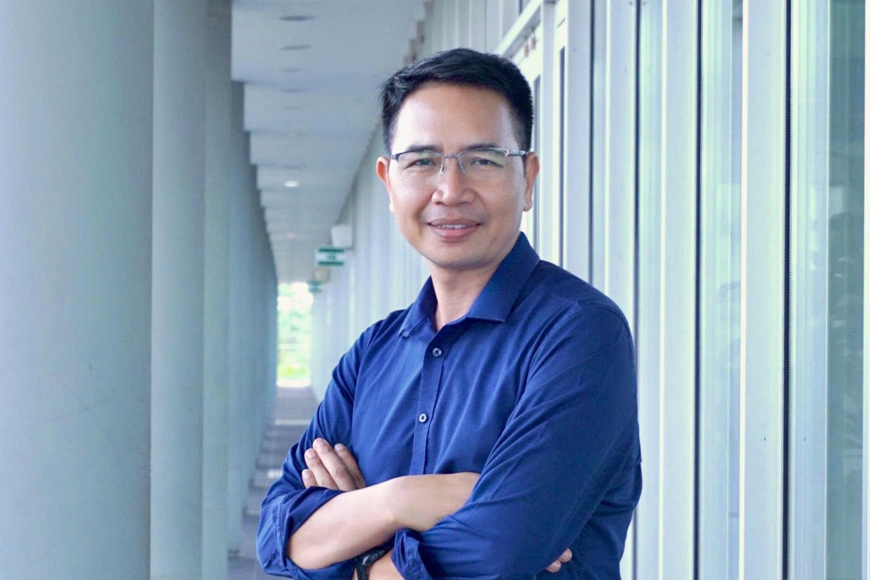 Cận cảnh một người đàn ông mặc áo sơ mi màu cam đang nhìn vào máy ảnh, mỉm cười trong một khung cảnh tươi sáng. 